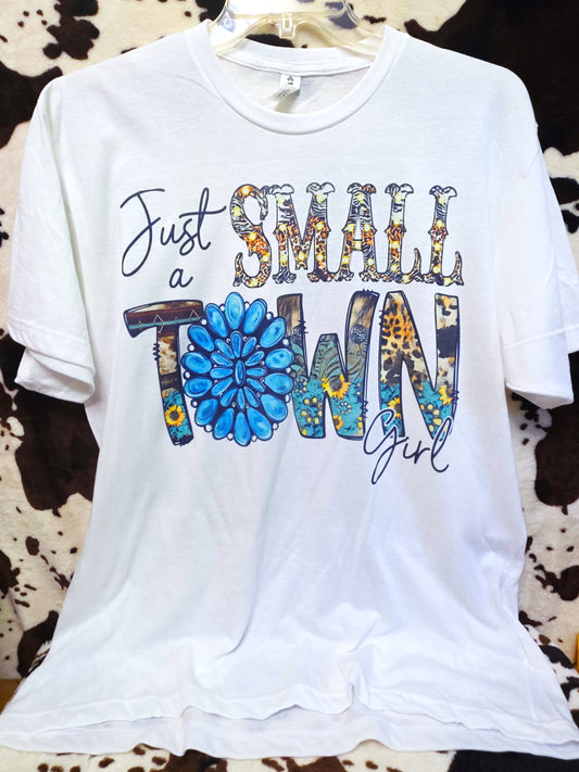 A white short-sleeve graphic T-shirt hanging on a clear hanger against a cowhide-patterned backdrop. The front graphic reads 'Just a SMALL Town Girl' with decorative floral and patterned lettering.
