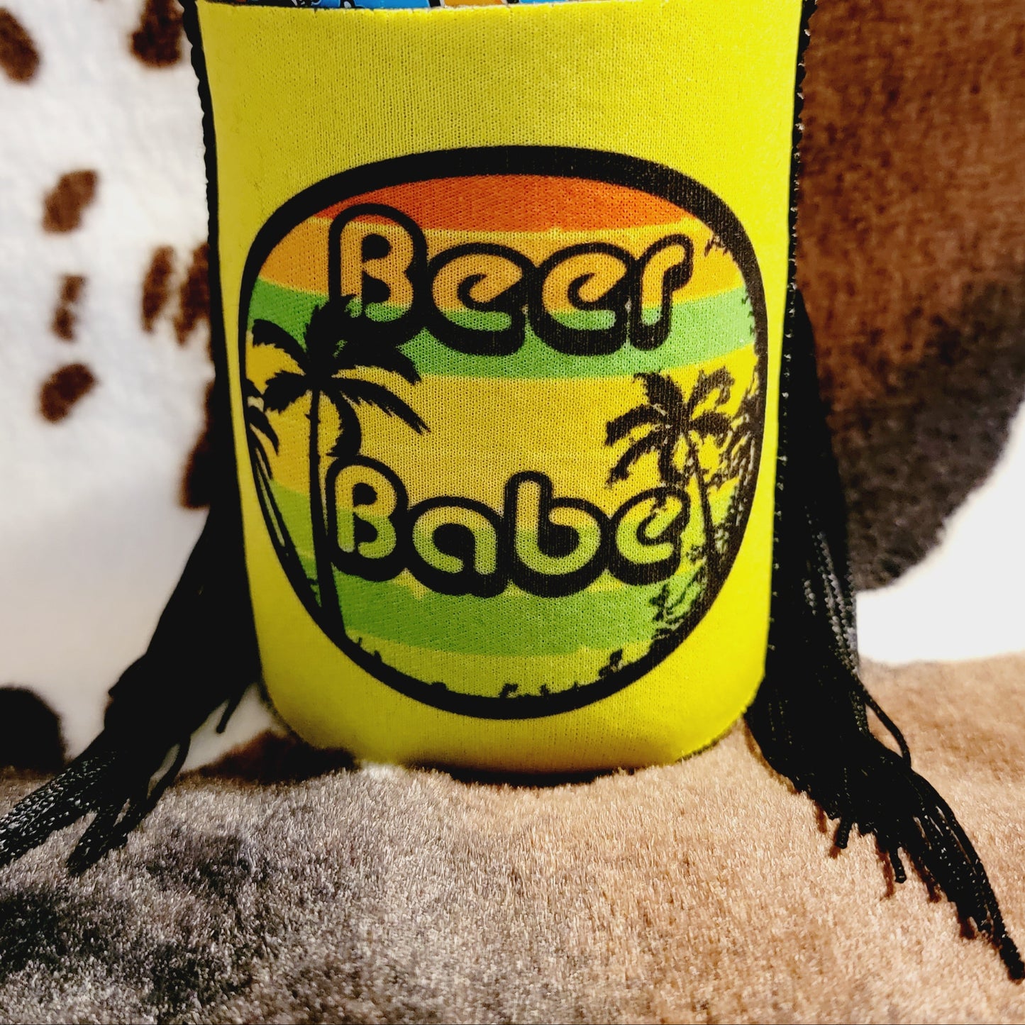 A neoprene can cooler koozie with a 'BEER BABE' text and design, predominantly yellow with green and orange accents, and a palm tree graphic.