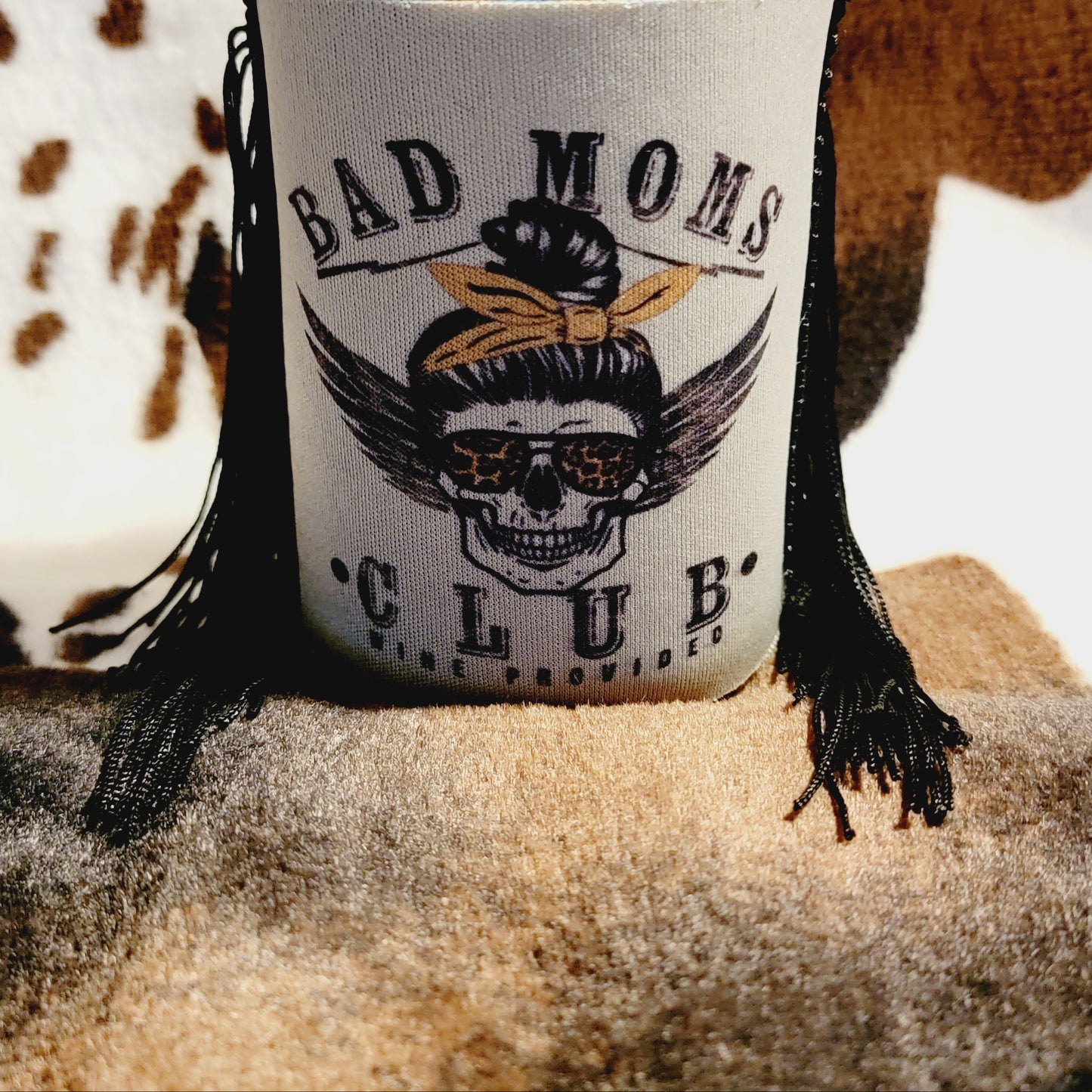 A neoprene can cooler koozie with 'BAD MOM'S CLUB' text and a skull design, holding a blue-canled beverage.