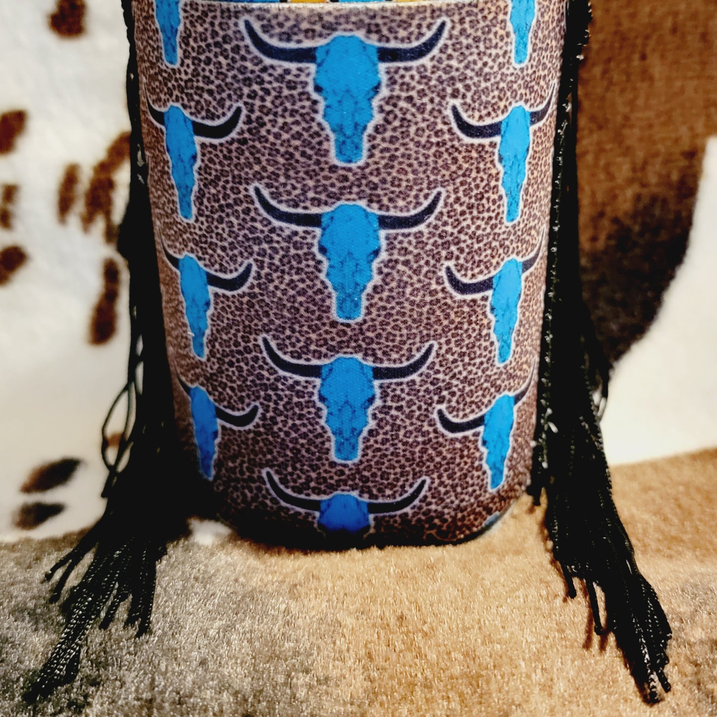 A neoprene drink sleeve with a western koozie design featuring a bull skull and leopard print patterns, predominantly blue with fringe detailing.