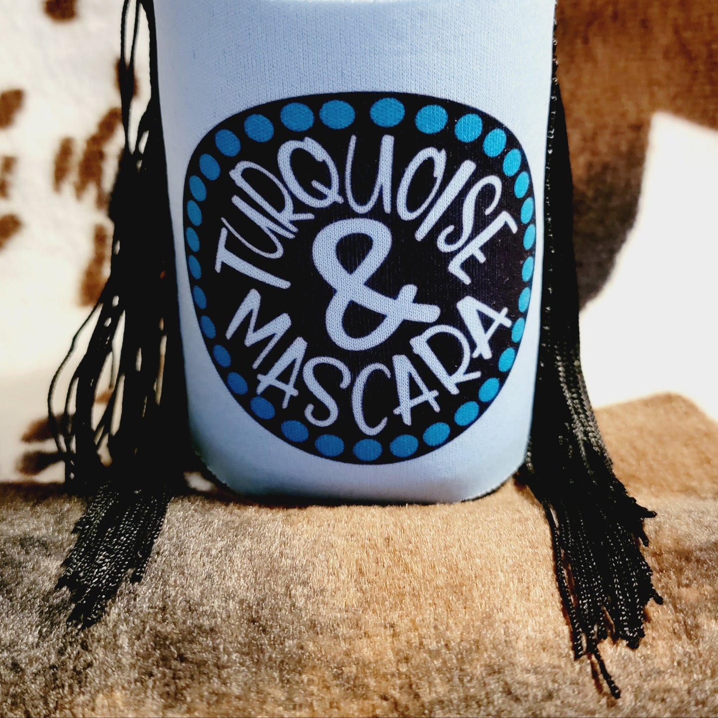 A turquoise blue neoprene koozie with the text 'Turquoise & Mascara' printed on it, holding a can.