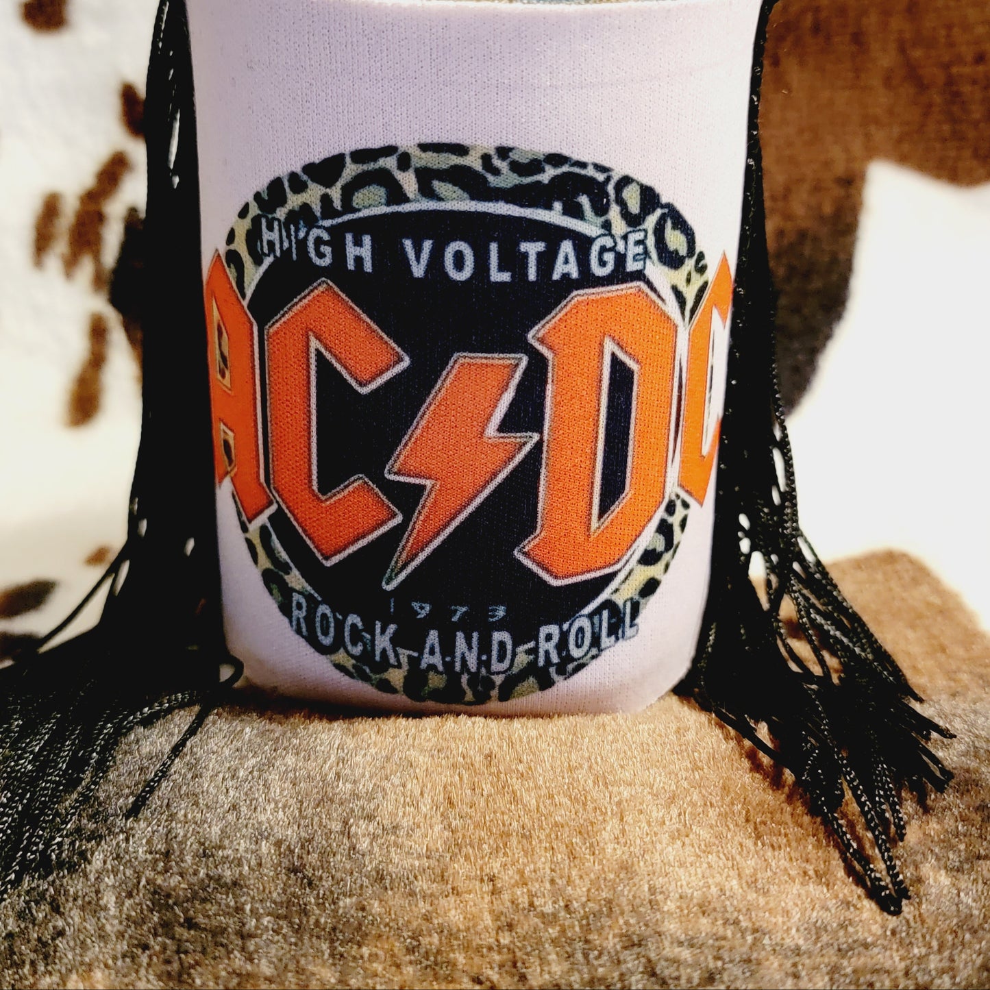 AC/DC themed koozie with a leopard print design, holding a can, against a cowboy hat and boots backdrop.